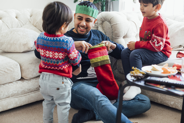 A mid-adult man sitting with his two sons in the home living room during the Christmas period time. The father is opening a stocking while the children watch.