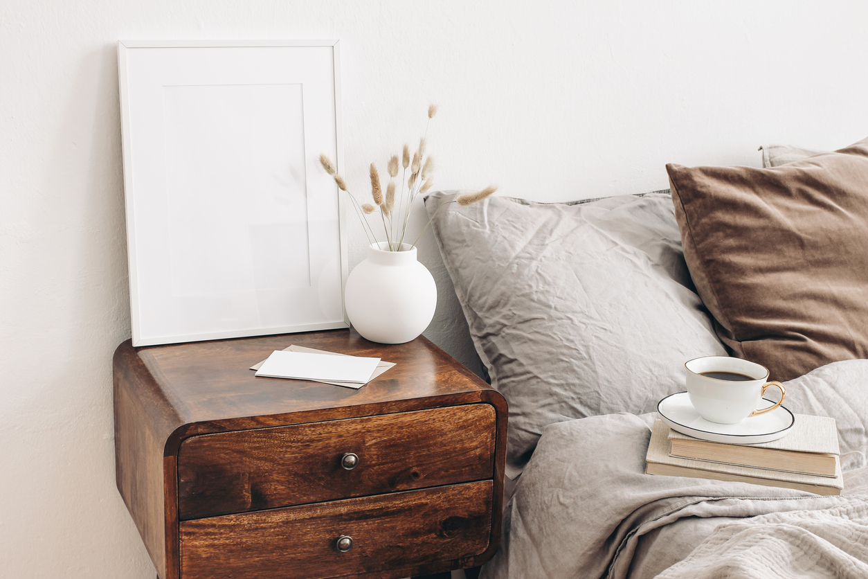 Portrait white frame mockup on retro wooden bedside table. Modern white ceramic vase, dry Lagurus ovatus grass. Cup of coffee and books in bed. Beige linen pillows in bedroom. Scandinavian interior.