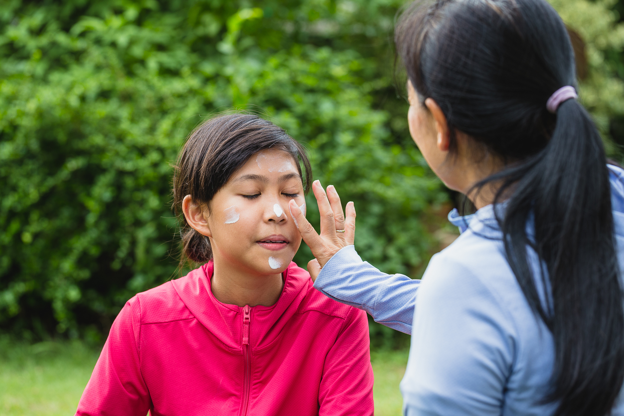 Asian mother applying sunscreen lotion to protect her daughter's face before exercise while setting on floor in the park at outdoors