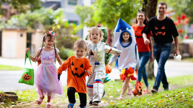 Child in Halloween costume. Mixed race Asian and Caucasian kids and parents trick or treat on street. Little boy and girl with pumpkin lantern and candy bucket. Baby in witch hat. Autumn holiday fun.