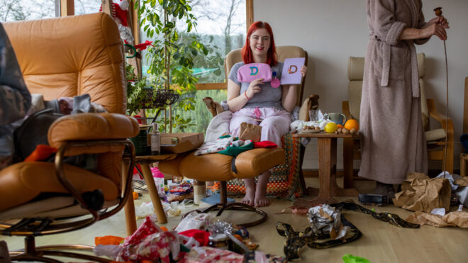 A teenager with vibrant dyed red hair with her grandmother in their living room in their home in Hexham, Northumberland in the North East of England. She is looking and smiling at the camera as she holds a new diary and pencil case which she has been gifted for Christmas.