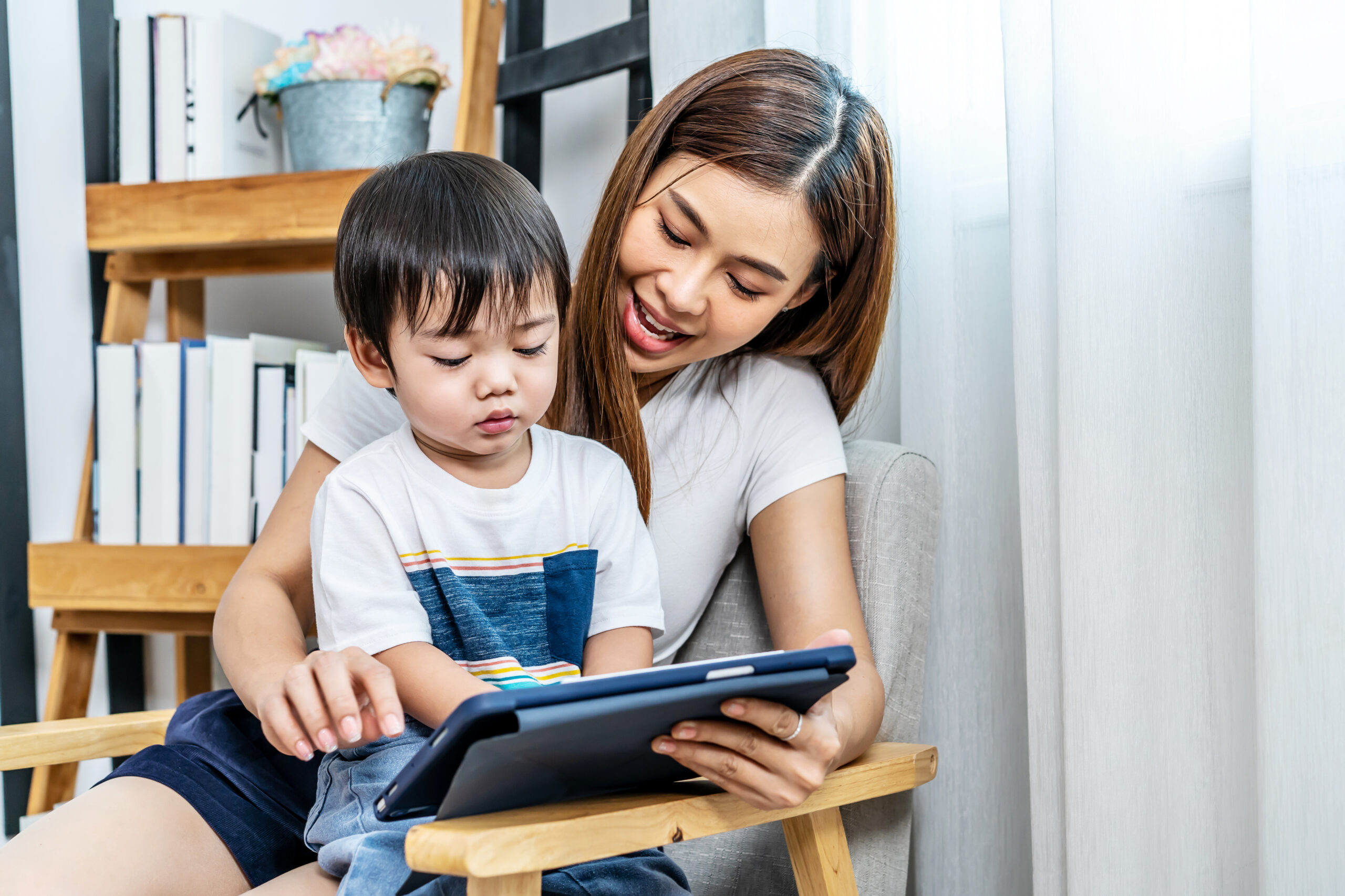 Young asian mother her little adorable son using tablet looking at  screen enjoying watching funny social media video doing online shopping relaxing on sofa at home together