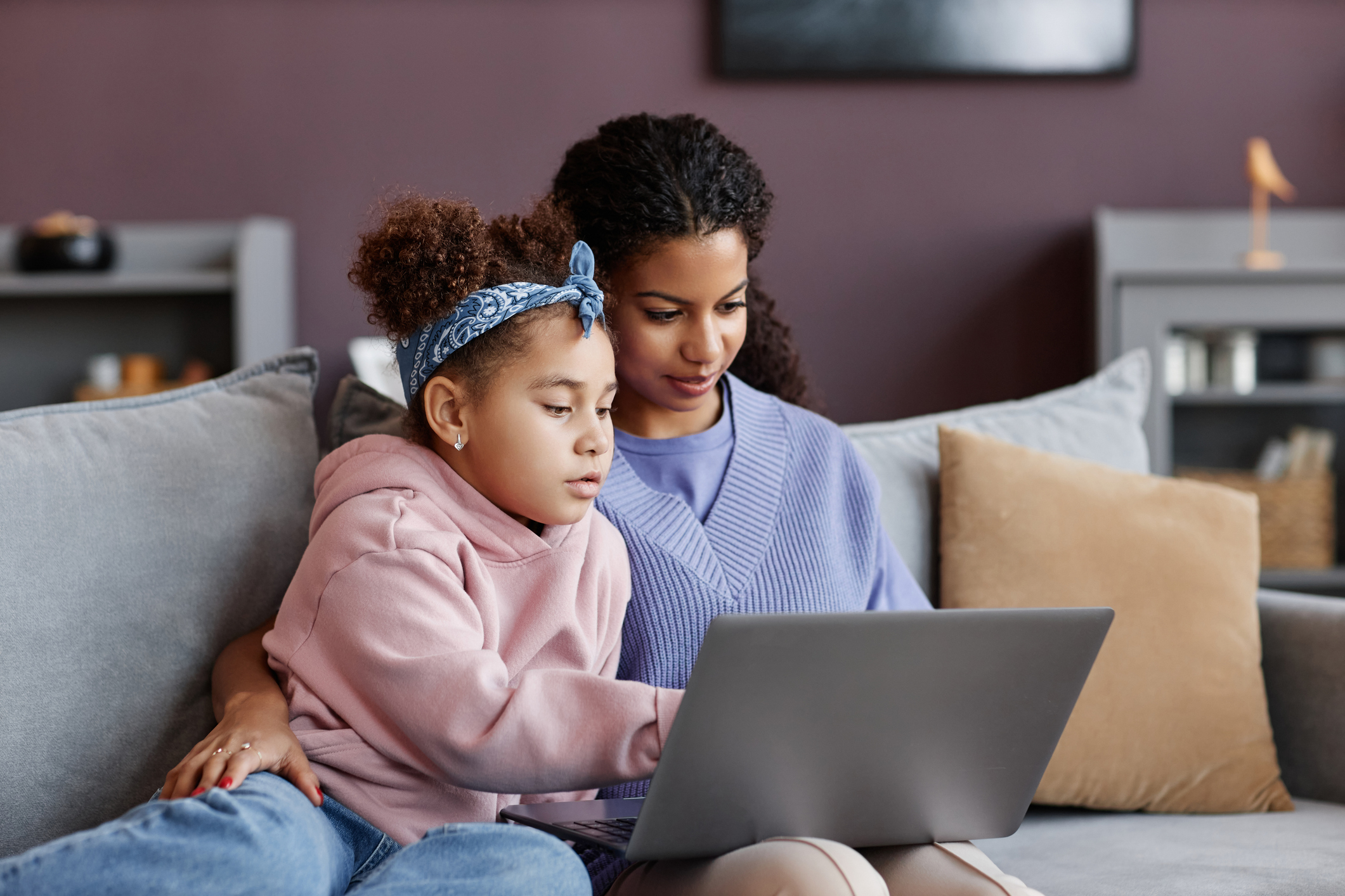 Black mother and daughter using laptop sitting on sofa together