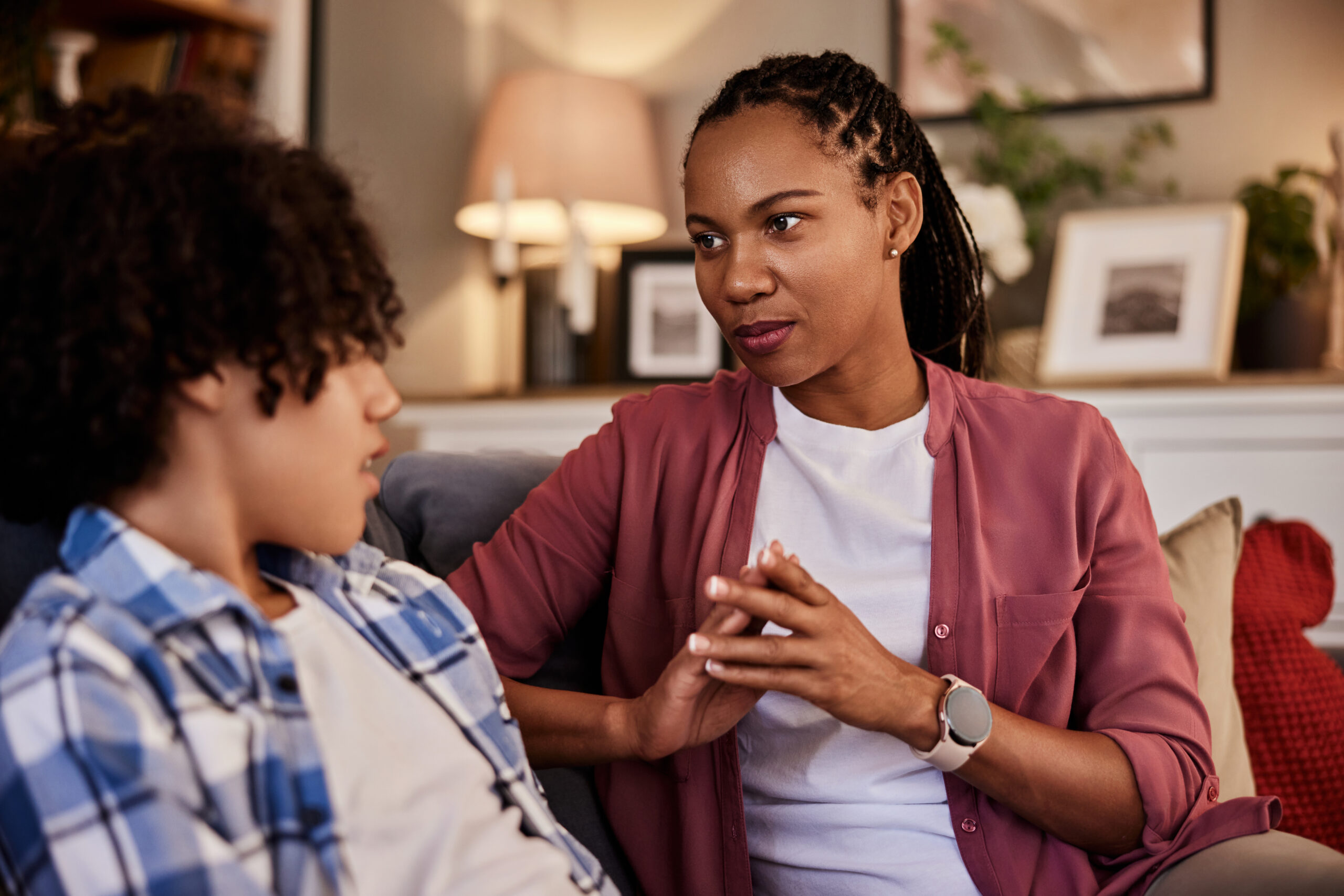 An African-American woman sitting on a couch with her teenage son and talking.