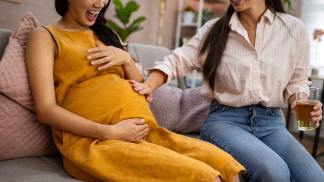 two women, one pregnant and wearing a yellow dress, and the other in a white shirt and jeans touching the other's baby bump