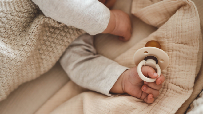 arms of a sleeping baby against a beige blanket. one hand clutching a pacifier