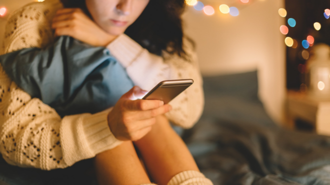 young woman on a bed in a white sweater clutching a pillow and looking at a phone with lights in the background